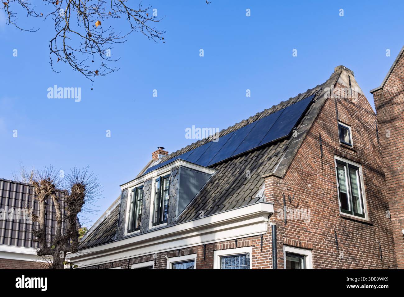 Pannelli solari installati sul tetto di mattonelle scure di un tradizionale e monumentale edificio olandese in mattoni contro un cielo blu brillante in una giornata di sole. Foto Stock