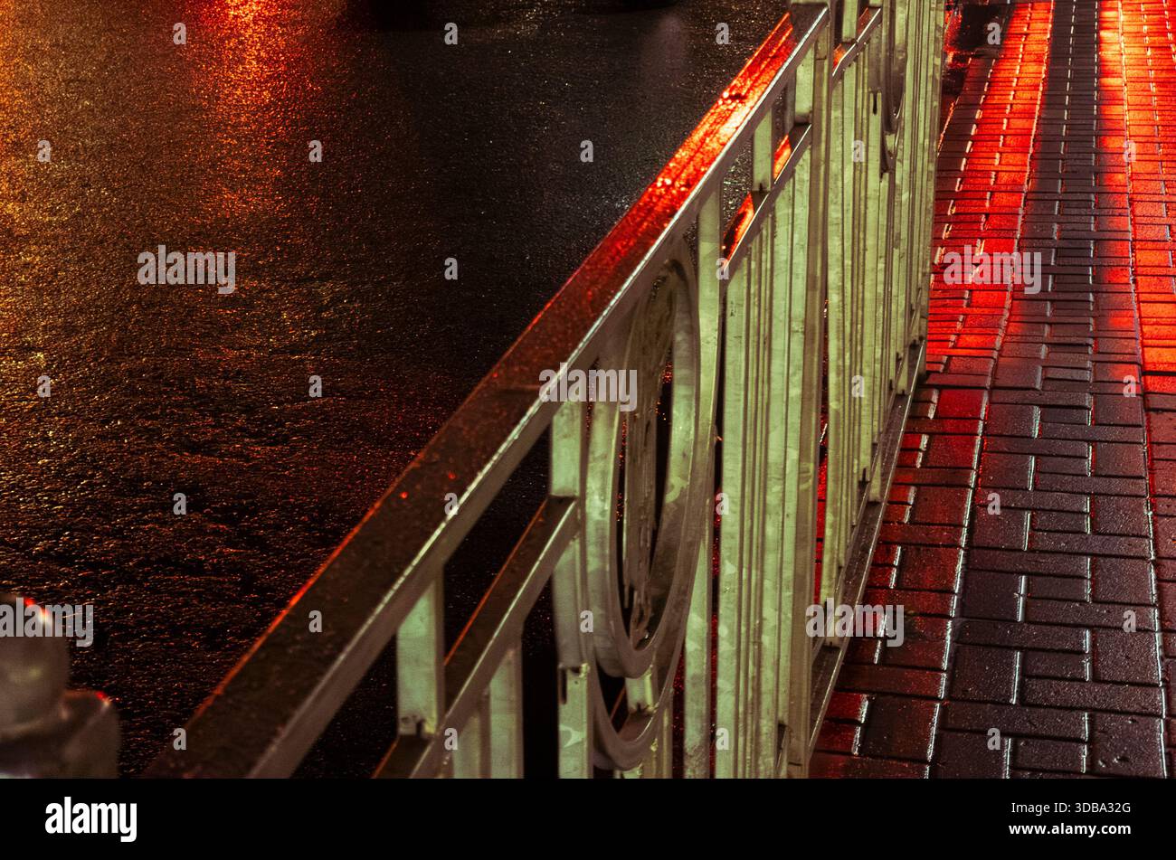 Pavimentazione urbana bagnata di notte che riflette la luce pubblica rossa lungo una ringhiera in metallo, marciapiede bagnato da pioggia soffusa con un bagliore vibrante, bokeh e su bagnato testurizzato Foto Stock