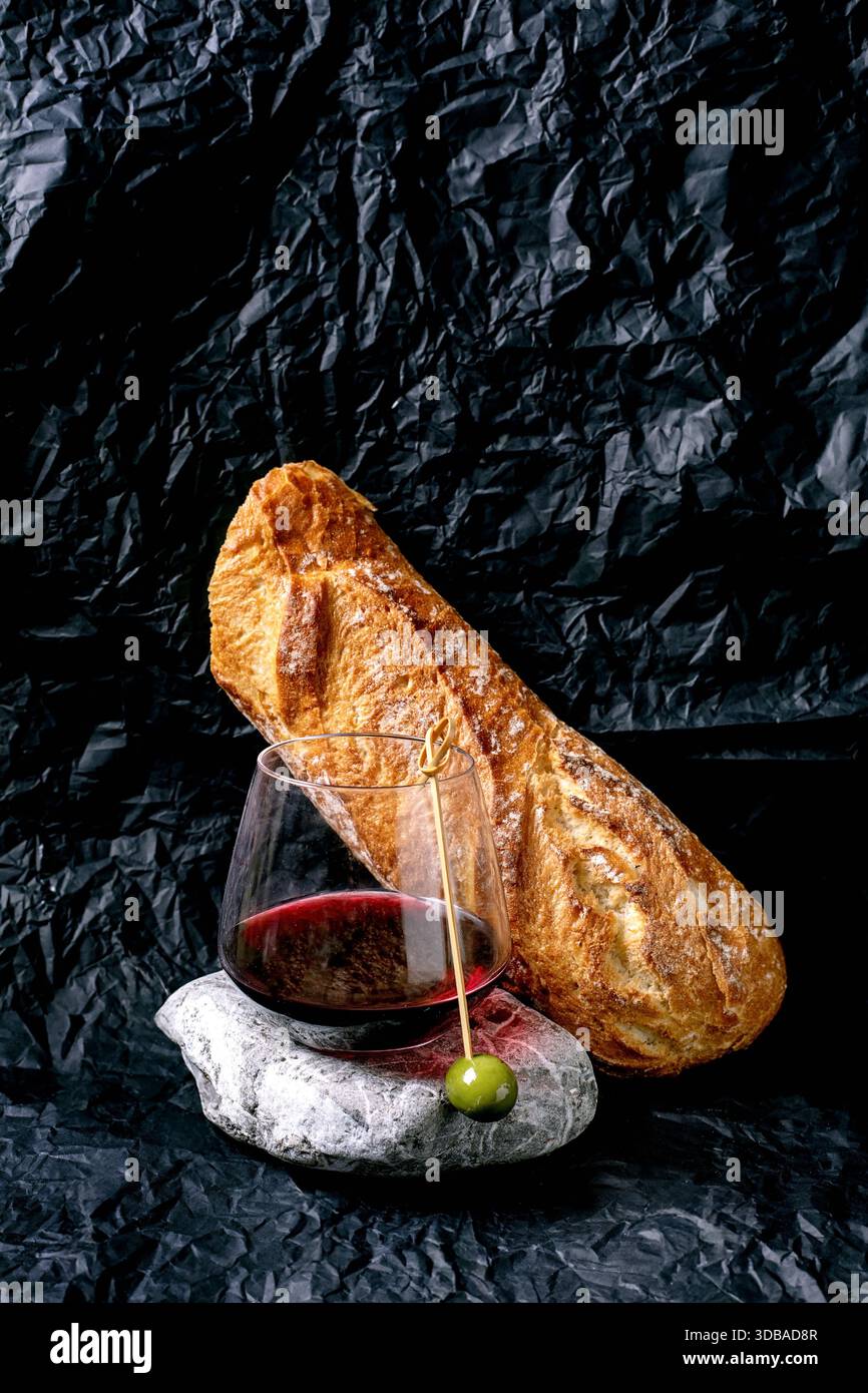 Natura morta con un bicchiere di vino rosso con un'unica asta di olive su pietra e pane bianco. Sfondo di carta nera arricciata. Concetto di pane e vino. Foto Stock