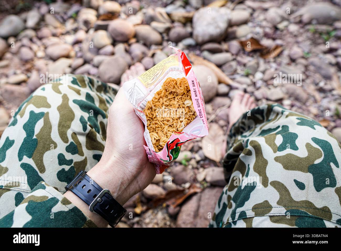 POV di una persona in pantaloni mimetici che tiene un pacchetto aperto di spaghetti istantanei asciutti mentre riposa su un terreno roccioso all'aperto. Foto Stock