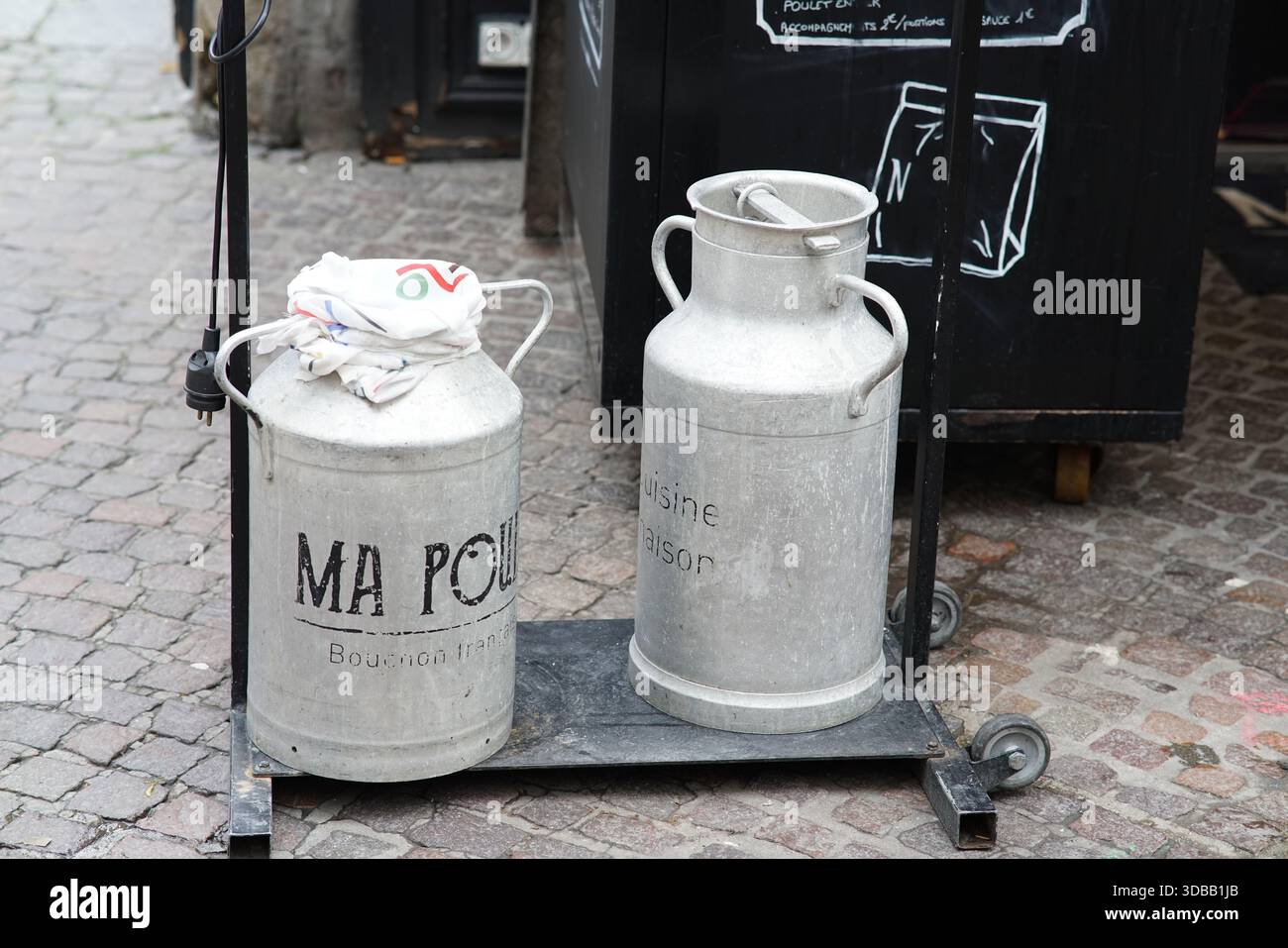 Due lattine di latte vintage in metallo esposte su uno stand con ruote all'esterno in una strada acciottolata. Nantes, Francia Foto Stock