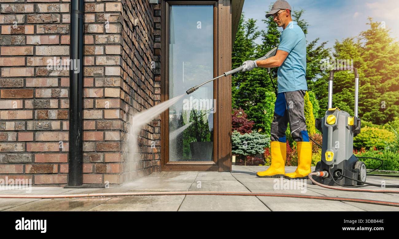 Una persona sta pulendo un muro di mattoni con un'idropulitrice fuori casa mentre si trova su un patio. Foto Stock