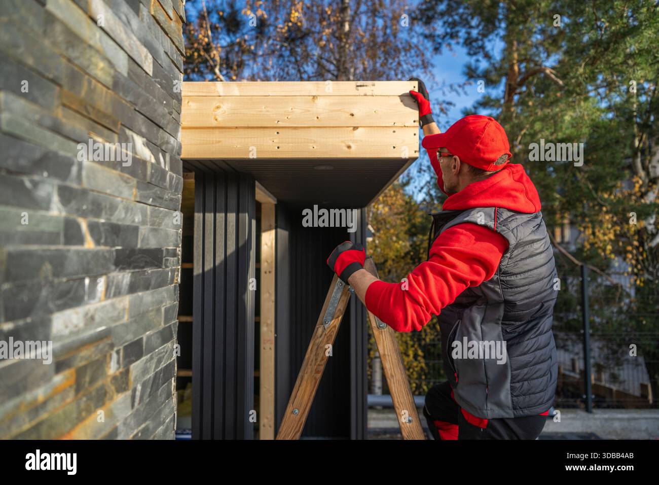 Una persona è su una scala, aggiungendo legno a una struttura vicino a una casa. Foto Stock