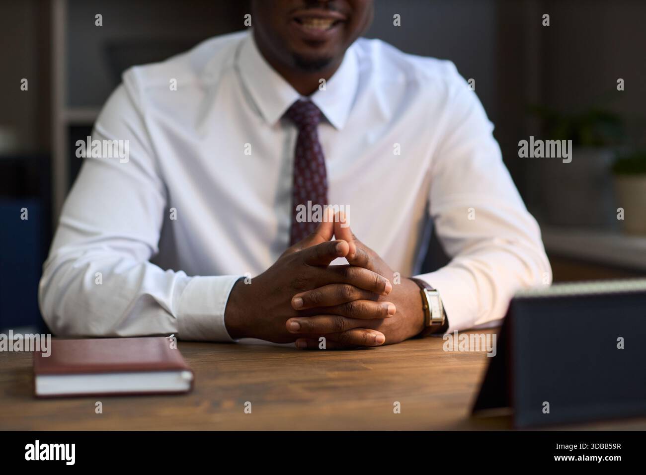 Uomo nero di mezza età seduto alla scrivania stringendo le mani e indossando camicia e cravatta formali, presentando una postura sicura in ufficio, suggerendo il preside della scuola al lavoro Foto Stock