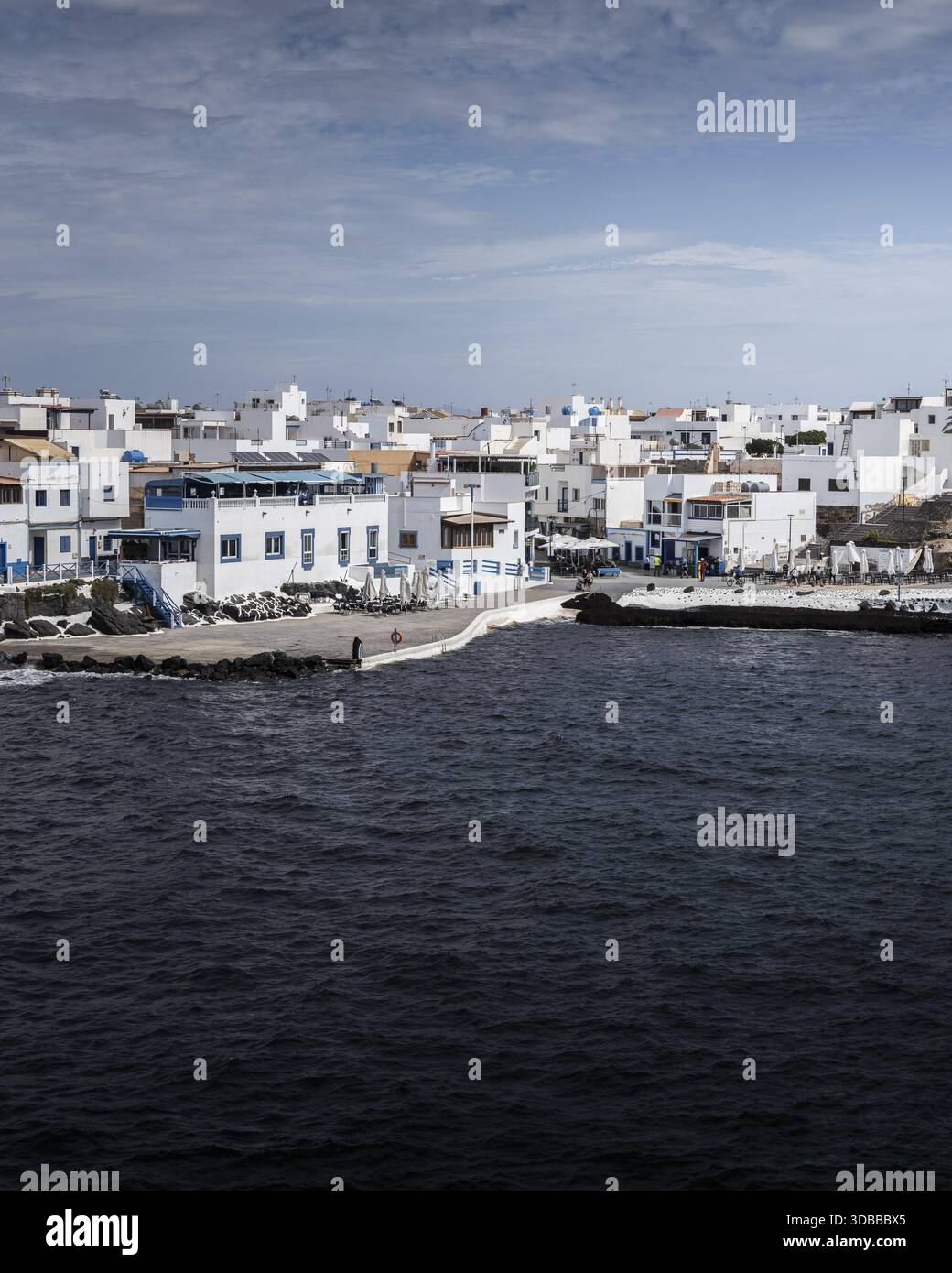 Vista degli edifici bianchi baciati dal sole con accenti blu che contrastano con il mare scuro e ondulato sotto un cielo parzialmente nuvoloso, Fuerteventura, Isole Canarie Foto Stock