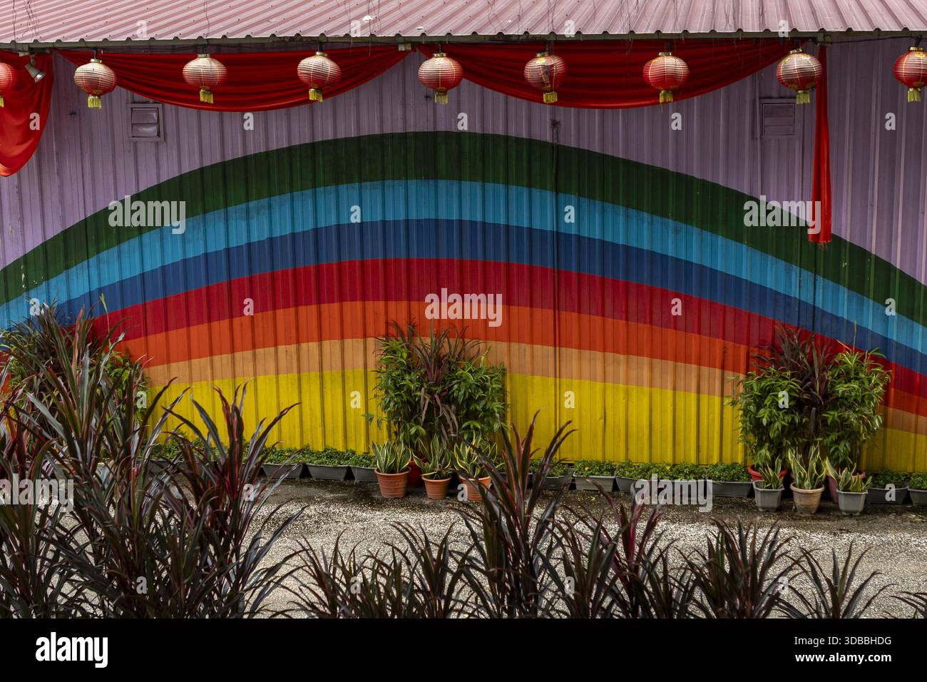 La vista dei vibranti arcobaleno irrompe attraverso una parete ondulata adornata da festose lanterne rosse, in contrasto con la lussureggiante vegetazione sottostante, Kuala Lumpur, Fed Foto Stock