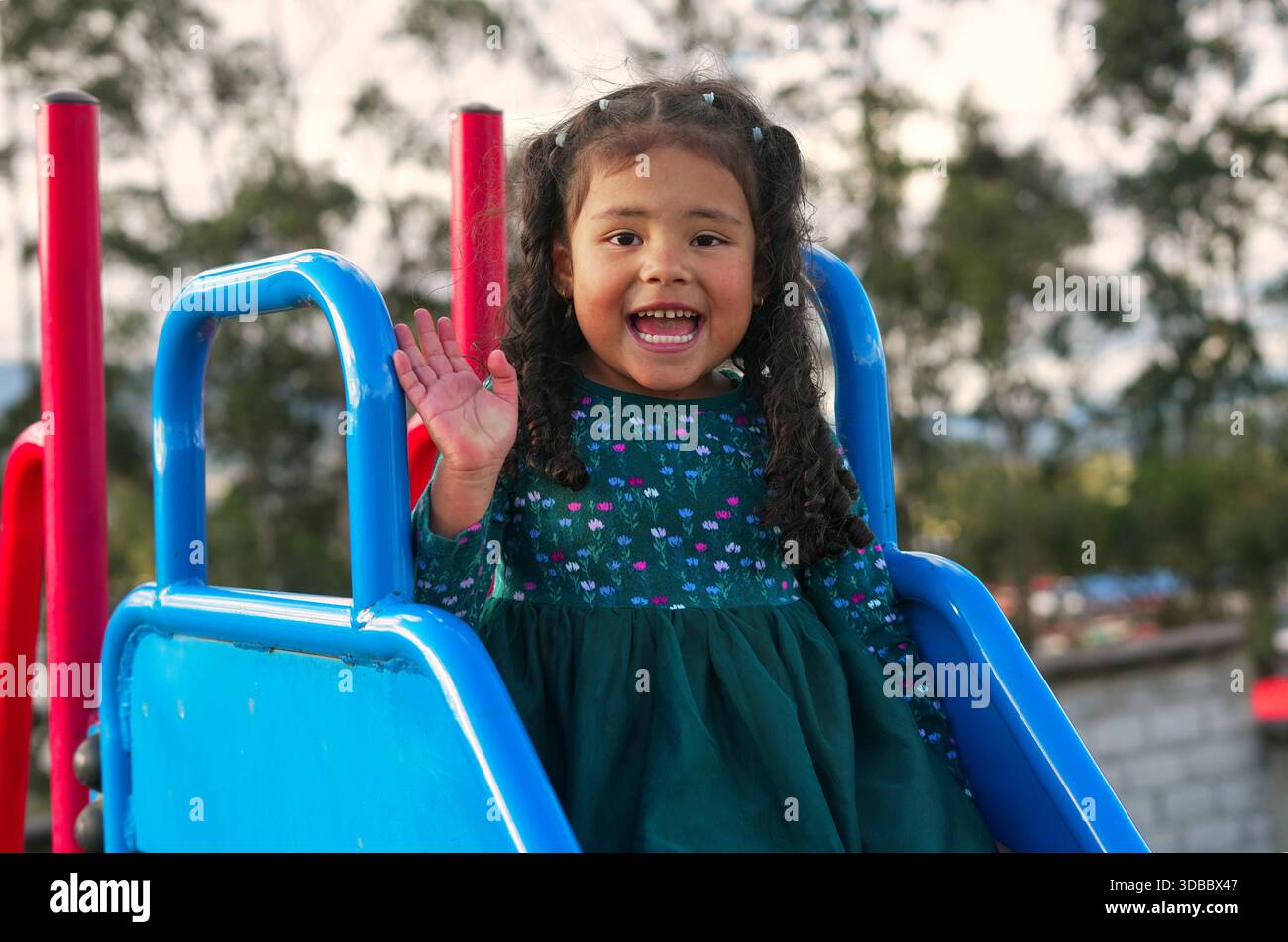 Sorridendo, in età prescolare, sventolando con gioia la mano mentre scivoli su uno scivolo blu vibrante in un parco giochi, per divertirsi all'aperto con una luminosa, S. Foto Stock