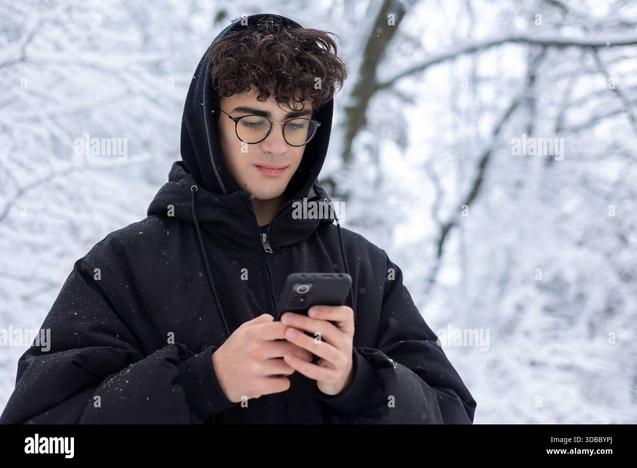 Ritratto di un teenager concentrato che usa lo smartphone all'aperto nello Snowy Winter Park. Giovane che controlla i social media o invia SMS, indossa occhiali e Black PUF Foto Stock