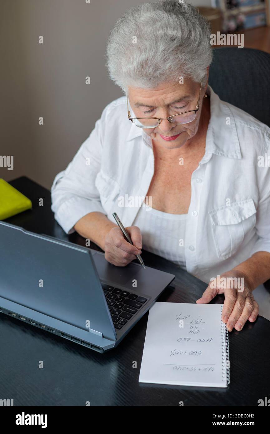 Donna anziana che utilizza un notebook a casa, paga bollette e gestisce le finanze online. Persone anziane che imparano le competenze digitali, fanno viaggi di pianificazione bancaria, Foto Stock