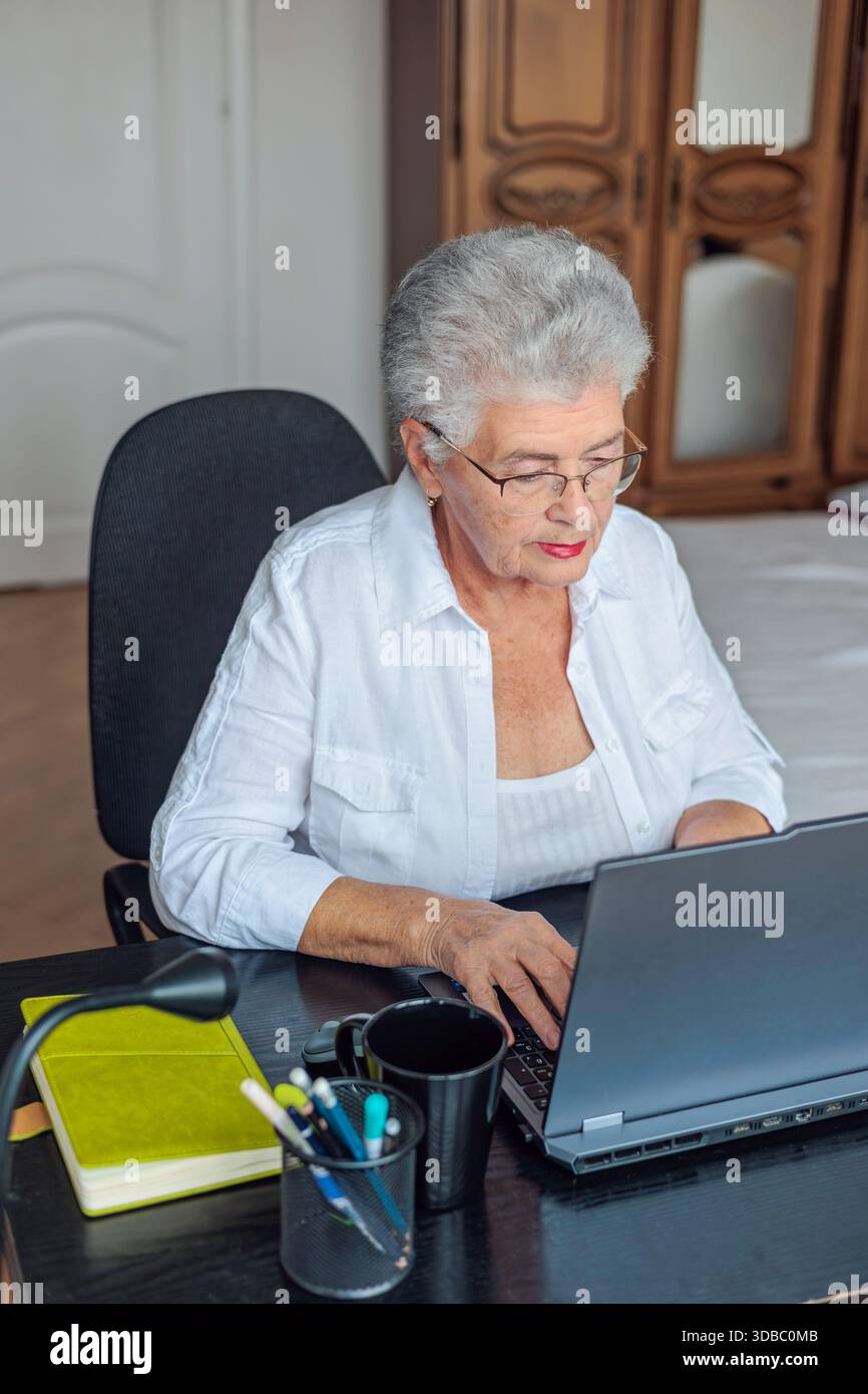 Donna anziana che utilizza un notebook a casa, paga bollette e gestisce le finanze online. Persone anziane che imparano le competenze digitali, fanno viaggi di pianificazione bancaria, Foto Stock