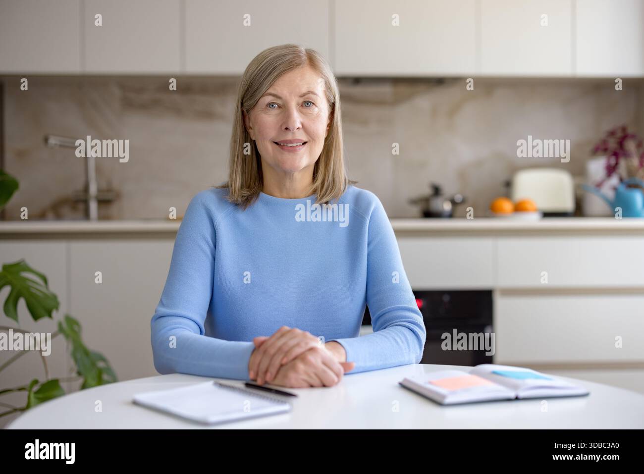 Donna matura seduta a un tavolo con i notebook, sorridendo con sicurezza alla fotocamera mentre lavora in remoto o partecipa a una videochiamata dalla sua moderna cucina domestica Foto Stock
