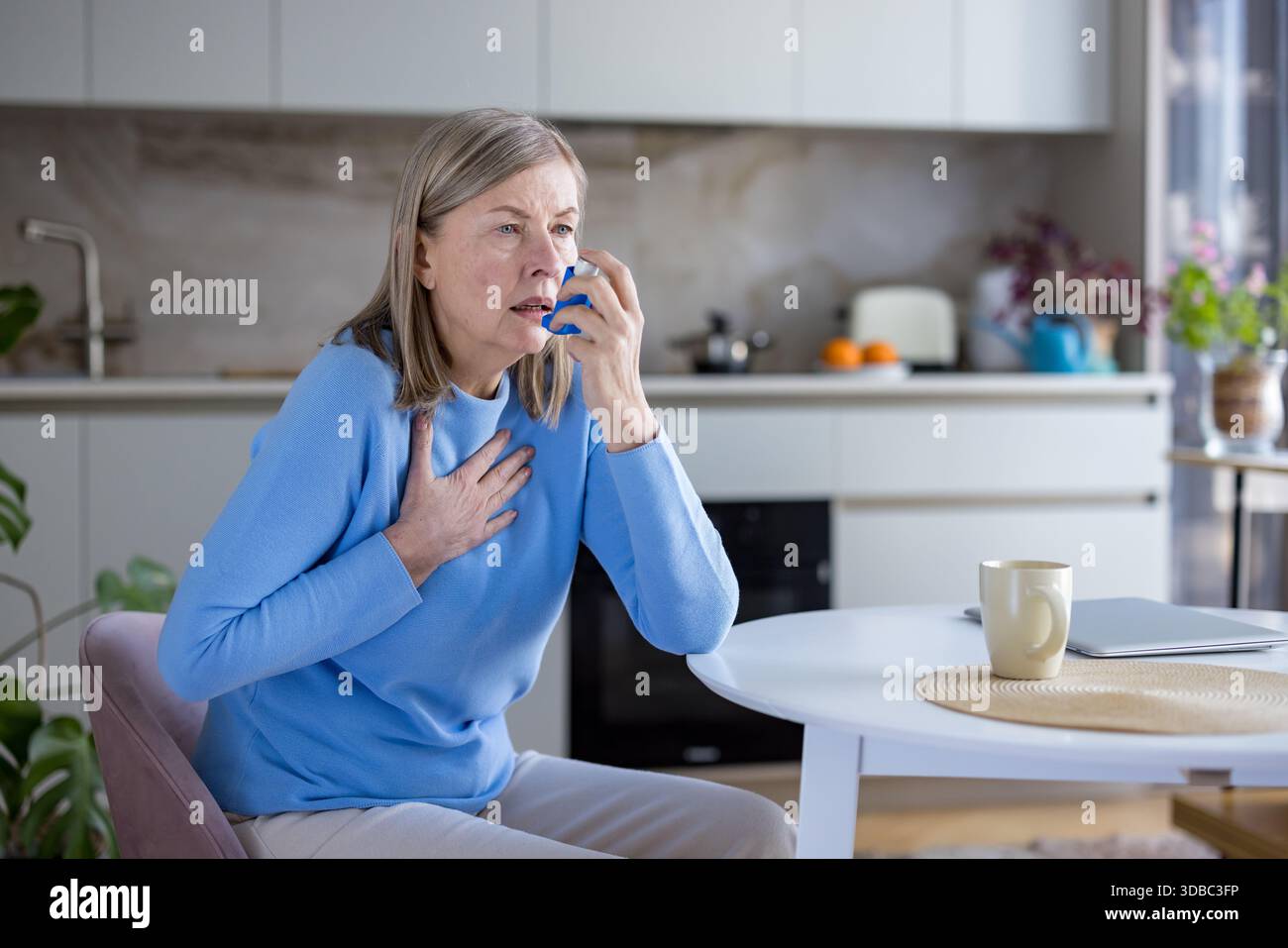 Donna anziana che soffre di un attacco d'asma e mancanza di respiro, somministrando urgentemente farmaci da un inalatore per alleviare le sue difficoltà respiratorie mentre è seduta in una moderna cucina domestica Foto Stock