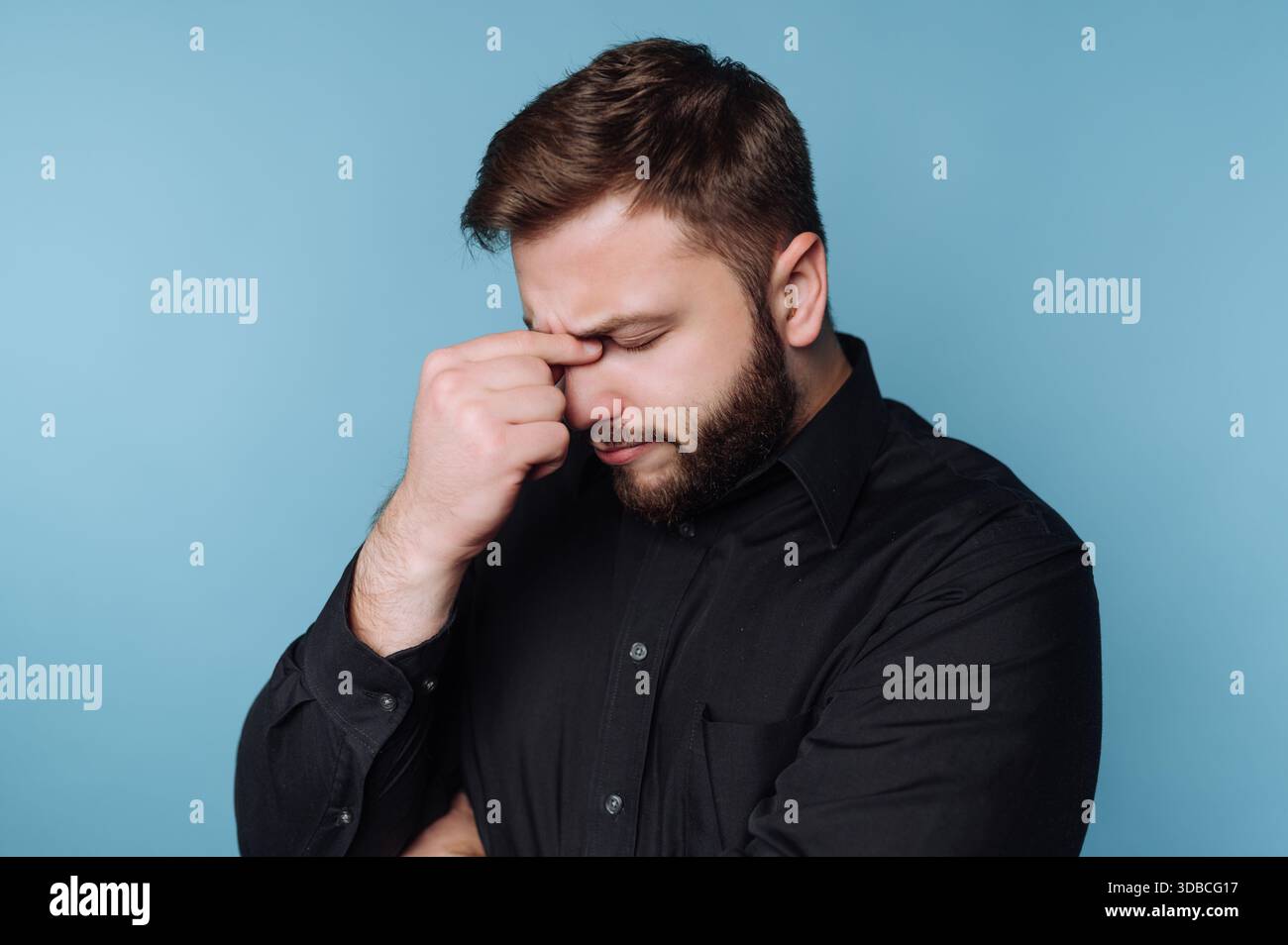 Un uomo sta in casa con uno sfondo blu. Tiene la fronte con una mano mentre guarda in basso. Sembra essere profondo nel pensiero o nel sentimento Foto Stock