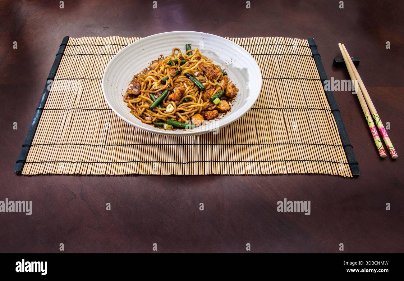 Una ciotola di spaghetti fritti al pollo serviti su un tappeto di bambù con bacchette, con verdure colorate e una presentazione di ispirazione asiatica. Foto Stock