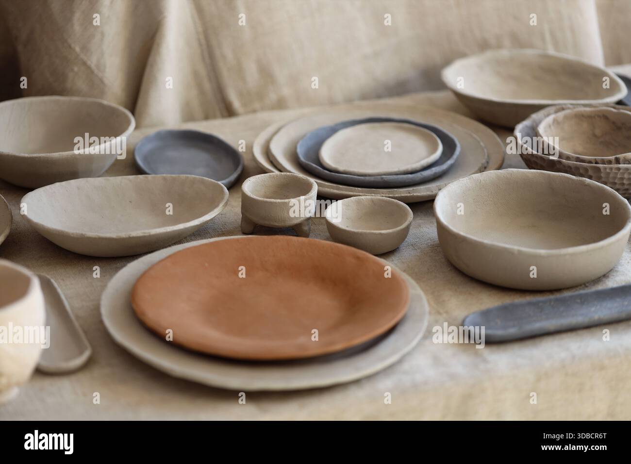 Assortimento di ciotole e piatti di argilla fatti a mano disposti su tessuto testurizzato. Fotografia di studio che mostra stoviglie in ceramica non smaltata con toni naturali e. Foto Stock