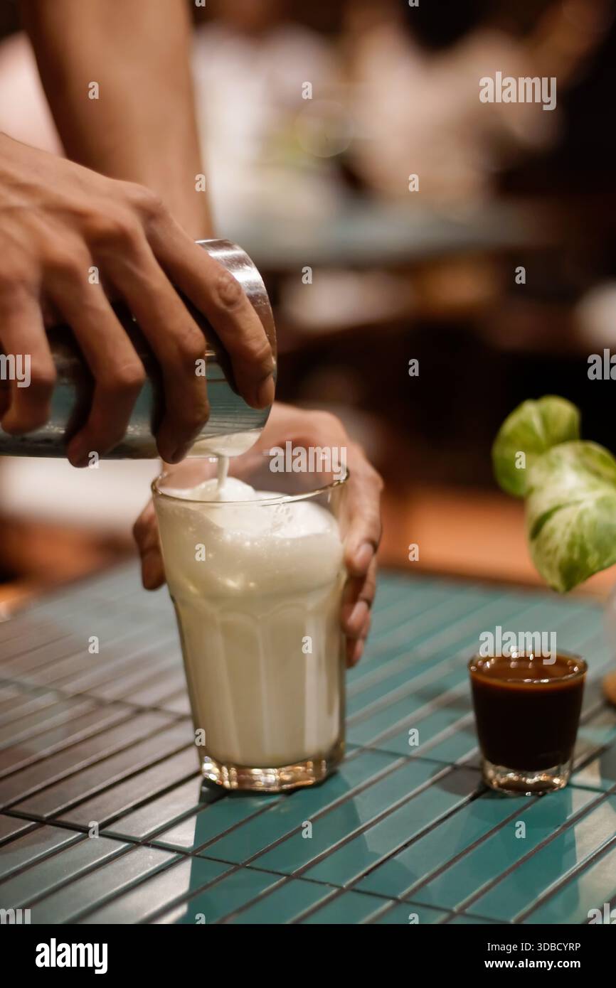 Il barista versa il latte montato nel bicchiere durante la preparazione del latte sul bancone del caffè, per chiudere il processo di preparazione del caffè Foto Stock