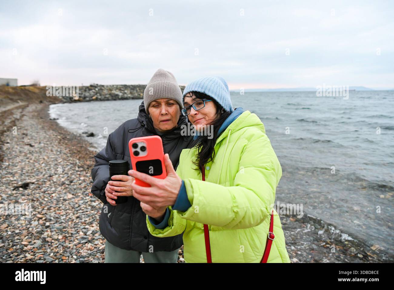 Due amici russi di 50 e 60 anni scattano un selfie durante una passeggiata primaverile al mare a Vladivostok, condividendo momenti di calma e un legame consapevole. Foto Stock
