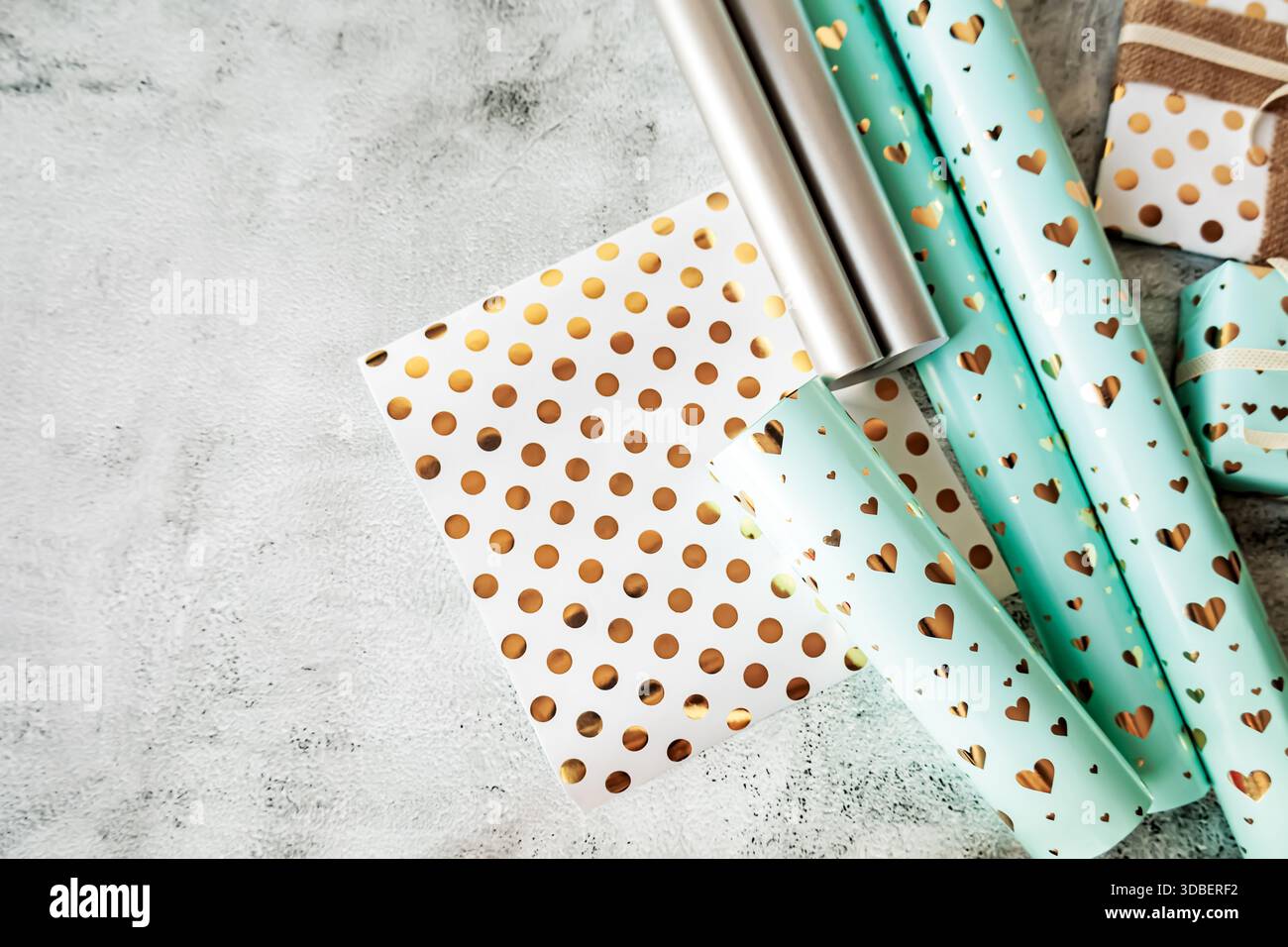 Disposizione piatta di rotoli di carta da regalo con motivi metallici dorati. Cuori turchesi, pois bianchi e tonalità argento su una superficie neutra testurizzata Foto Stock