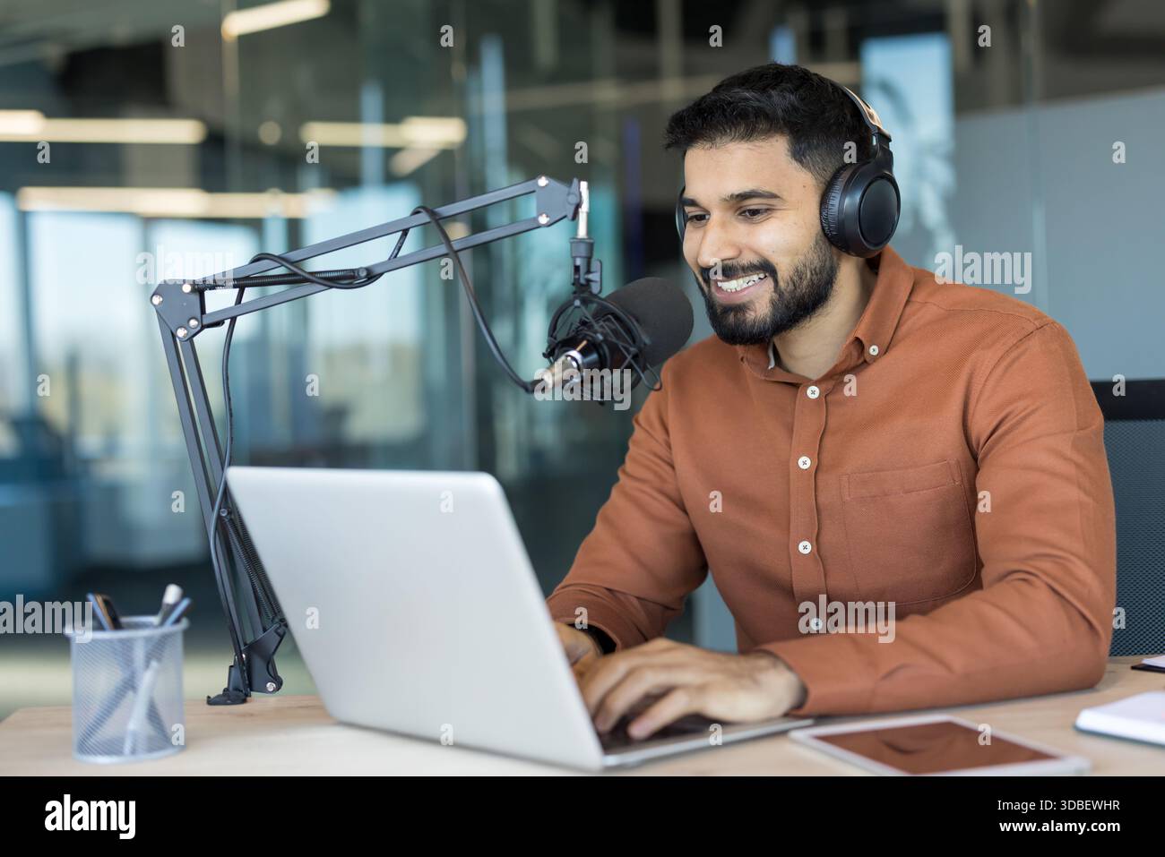 Giovane creatore di contenuti maschile che indossa le cuffie e parla in un microfono professionale mentre digita su un laptop, trasmettendo in diretta da uno studio moderno Foto Stock