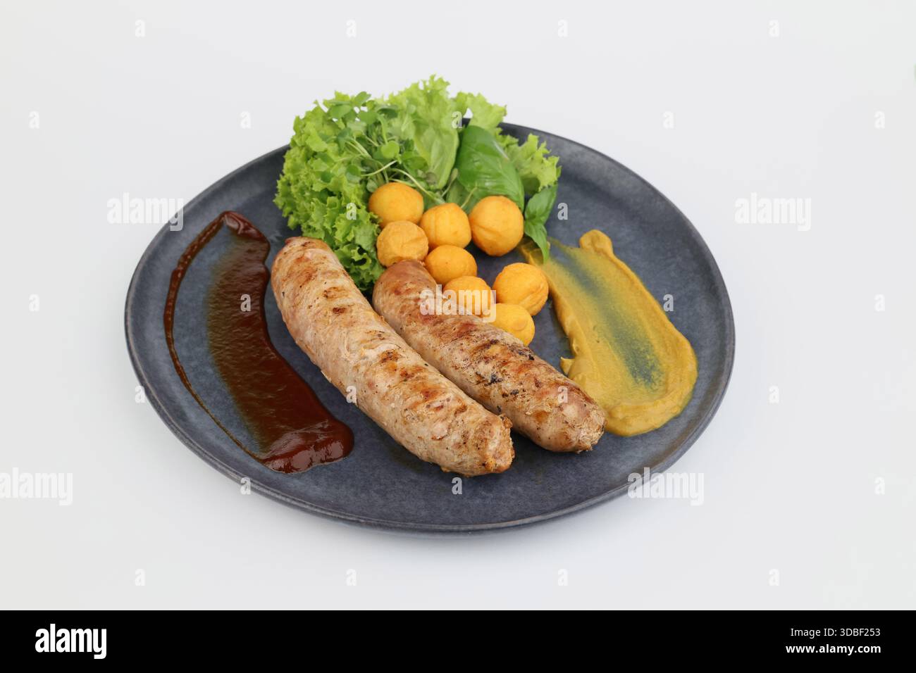 Salsicce grigliate con lattuga verde e crocchette di patate su un piatto scuro su sfondo bianco. Foto Stock