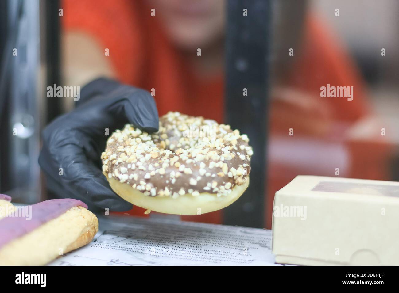 Concentrati sulle dita con i guanti neri che afferrano la ciambella cosparsa con un personaggio femminile sfocato nel maglione arancione alle spalle Foto Stock