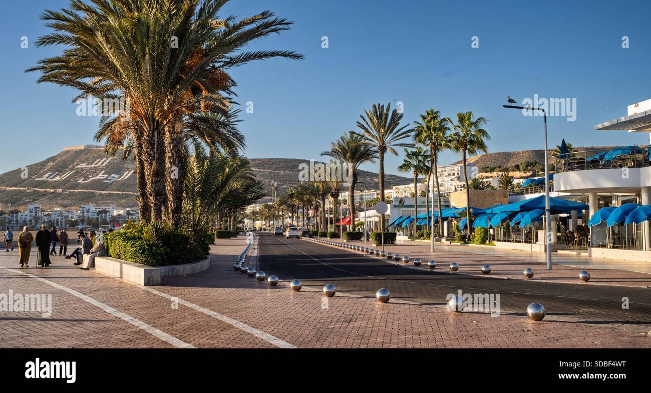 Passeggiata costiera soleggiata costeggiata da palme, ombrelloni blu e caffetterie all'aperto. Una tranquilla e invitante scena di strada con pedoni e servizi moderni a. Foto Stock