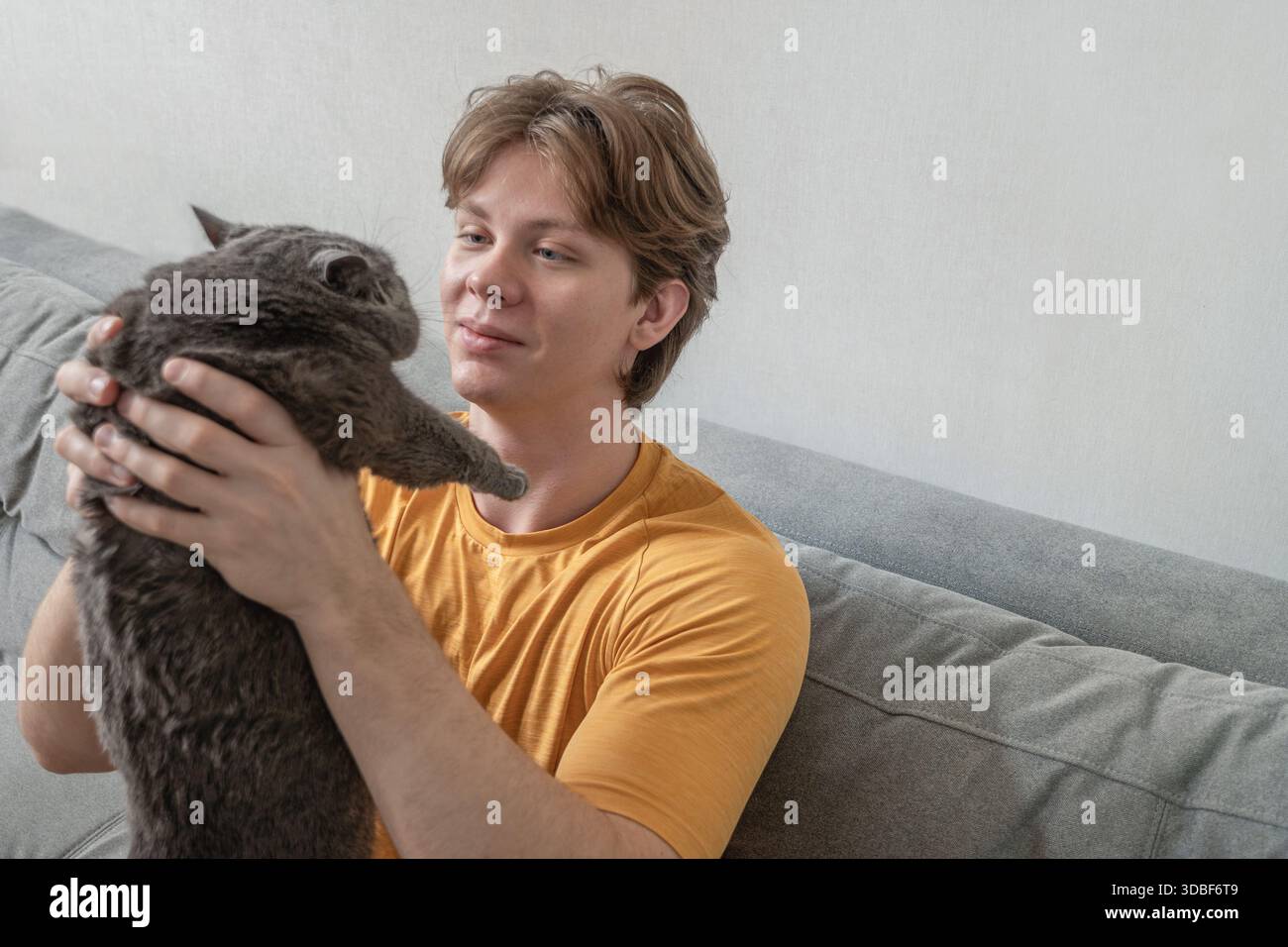Tenendo in mano il gatto e guardandolo con cura, il giovane passa del tempo tranquillo con il suo animale domestico a casa sul divano. Uomo che passa del tempo con il suo gatto a casa Foto Stock