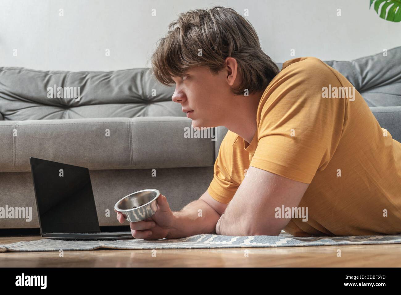 Lavorando da casa mentre si è sdraiati sul pavimento con un computer portatile, il giovane uomo trascorre del tempo con il suo gatto britannico in un accogliente soggiorno. Uomo che passa del tempo con il suo gatto a casa Foto Stock