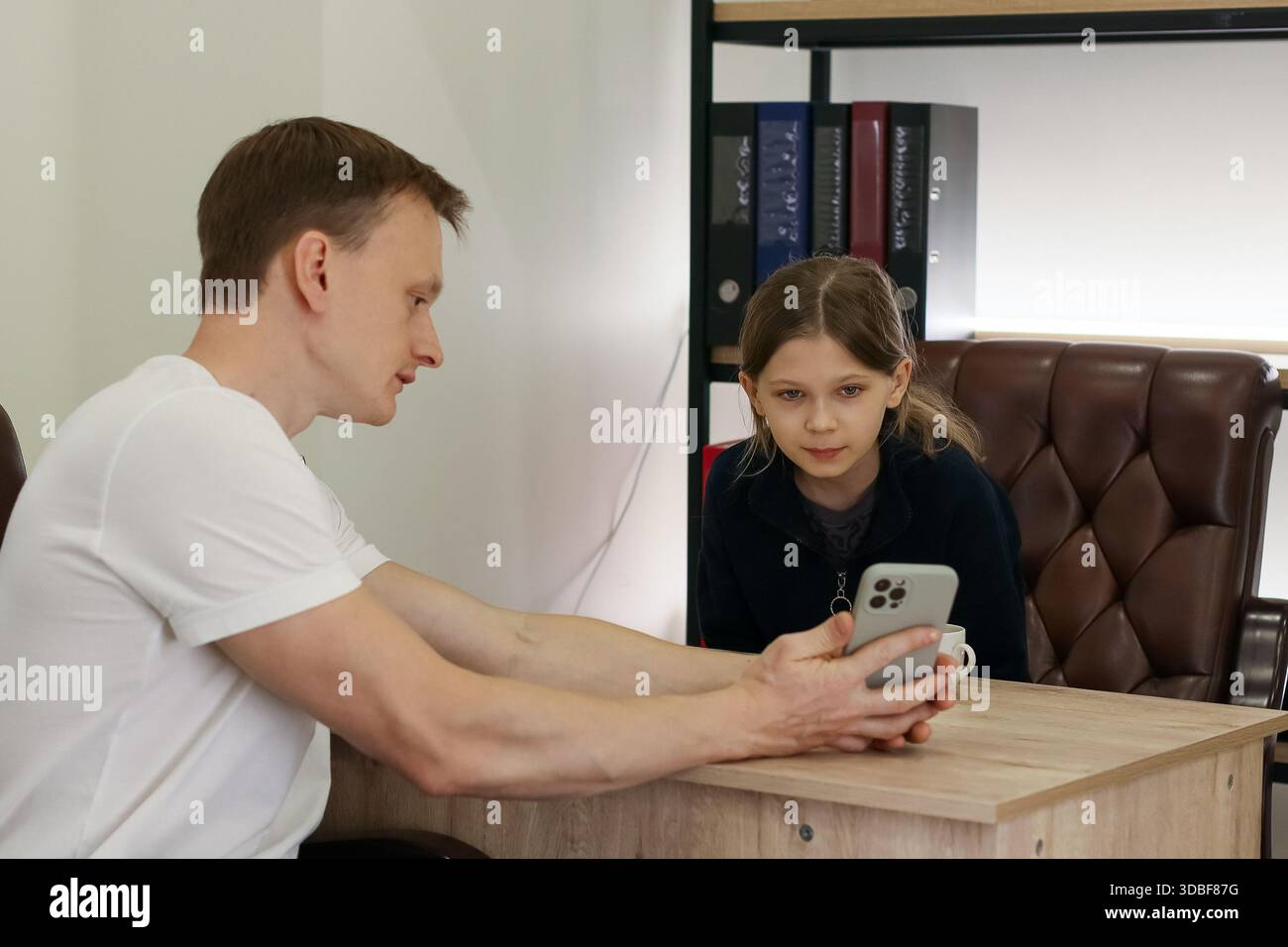Un maschio adulto si appoggia al tavolo dimostrando il dispositivo mobile al bambino seduto in un ambiente professionale con leganti e sedie Foto Stock