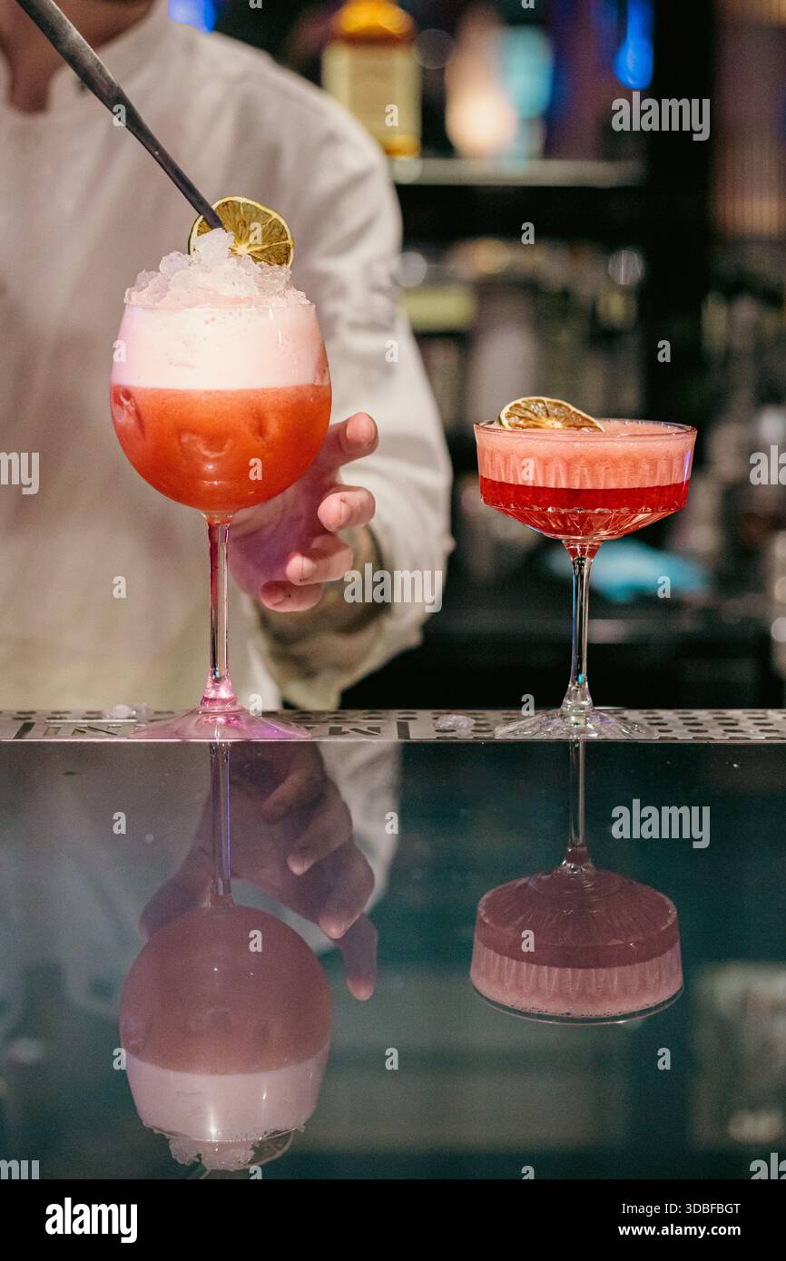 Elegante primo piano di due cocktail rossi in bicchieri a stelo su un moderno bancone da bar, caratterizzato da schiuma, guarnizione di agrumi e un'atmosfera vivace e notturna Foto Stock
