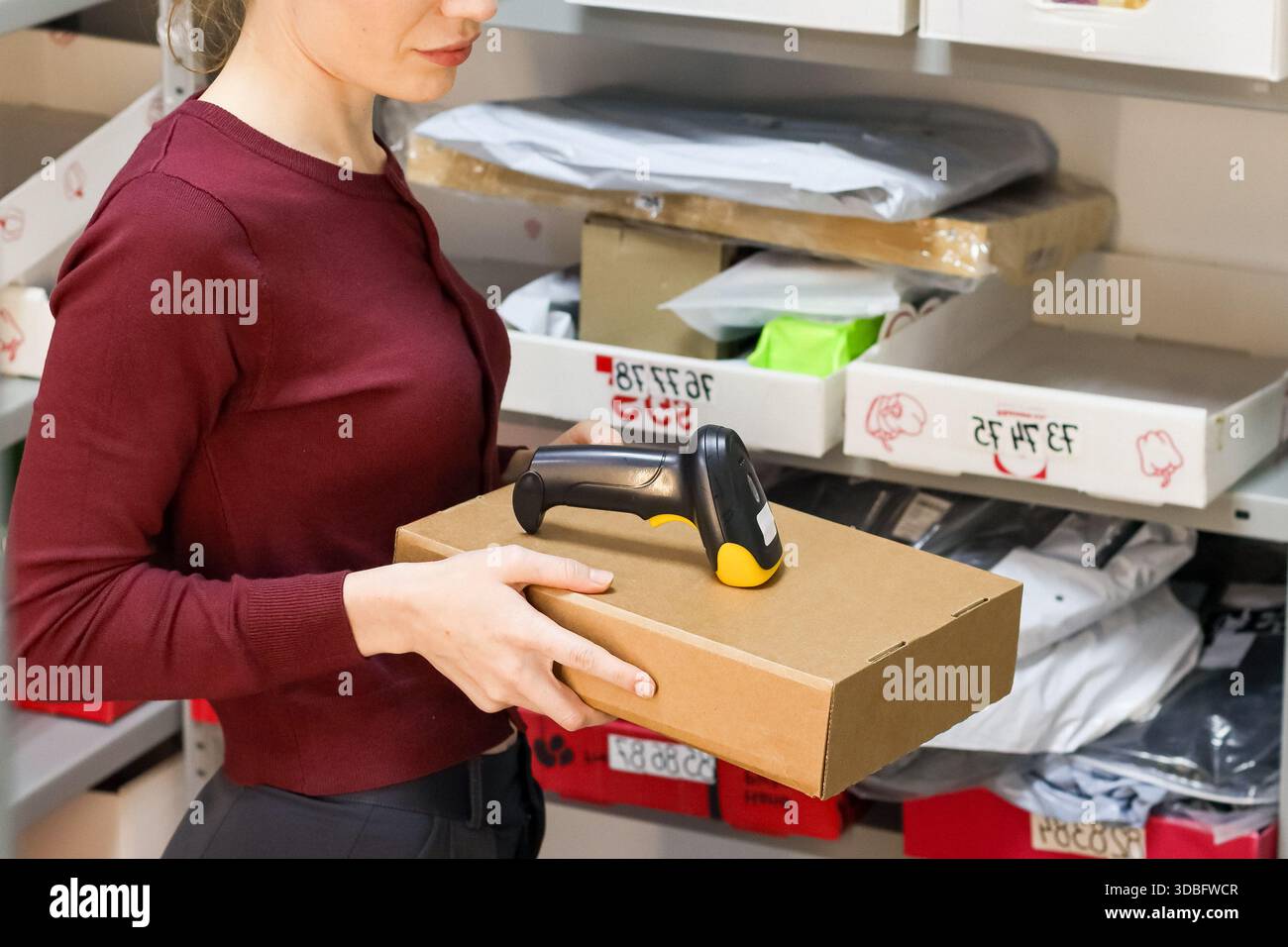 Una donna in camicia marrone inclina una scatola di cartone marrone e uno scanner di codici a barre con punta gialla nelle sue mani, stando accanto a scaffali metallici con confezioni. Foto Stock