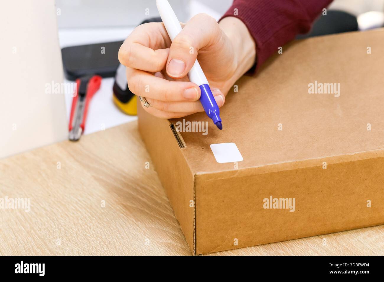 Immagine ravvicinata delle dita con la penna viola che scrive sull'etichetta bianca della confezione marrone in mezzo alle attrezzature. Foto Stock