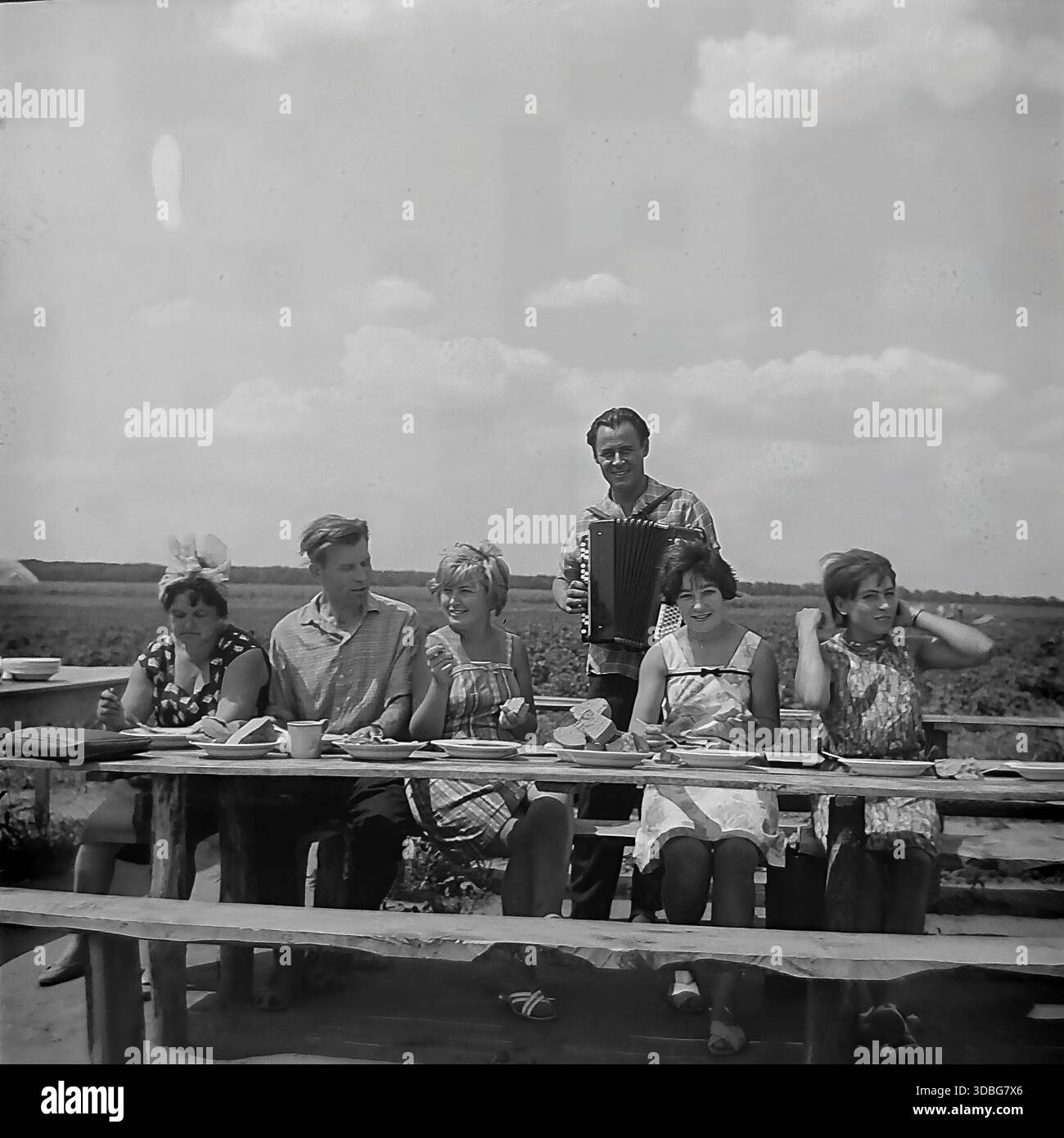 Foto d'archivio in bianco e nero che cattura un'allegra pausa pranzo in una fattoria collettiva nella regione di Donetsk, Ucraina, Donbas, anni '1960 I lavoratori e i cantanti in visita si siedono a un tavolo di legno grezzo in un campo aperto, mangiando zuppe e pane fresco, mentre un suonatore di fisarmonica sta dietro di loro fornendo intrattenimento musicale. Una vivida scena della vita rurale sovietica, della cultura "agitBrigade", dell'ospitalità del tempo e dell'atmosfera gioiosa della tranquilla campagna prima della guerra. Foto Stock