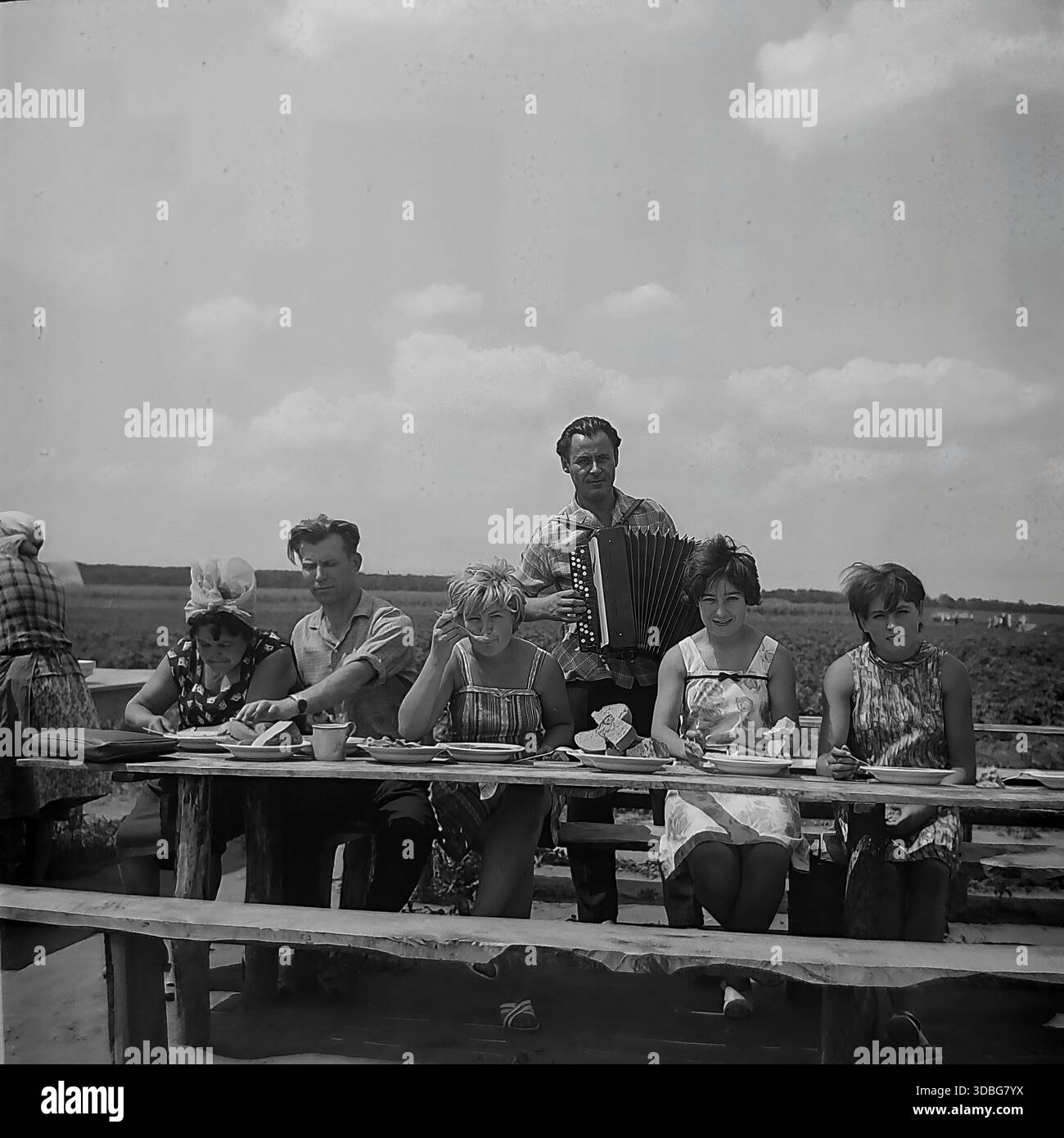 Foto d'archivio in bianco e nero che cattura un'allegra pausa pranzo in una fattoria collettiva nella regione di Donetsk, Ucraina, Donbas, anni '1960 I lavoratori e i cantanti in visita si siedono a un tavolo di legno grezzo in un campo aperto, mangiando zuppe e pane fresco, mentre un suonatore di fisarmonica sta dietro di loro fornendo intrattenimento musicale. Una vivida scena della vita rurale sovietica, della cultura "agitBrigade", dell'ospitalità del tempo e dell'atmosfera gioiosa della tranquilla campagna prima della guerra. Foto Stock
