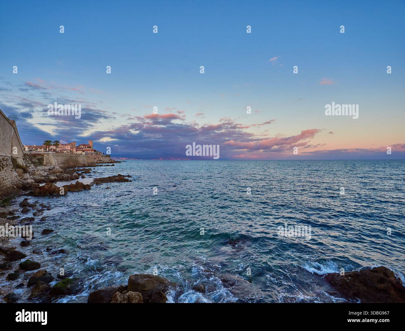 Tramonto costiero sul mare calmo con le storiche mura di pietra e la costa rocciosa con la città costiera di Antibes, Francia Foto Stock