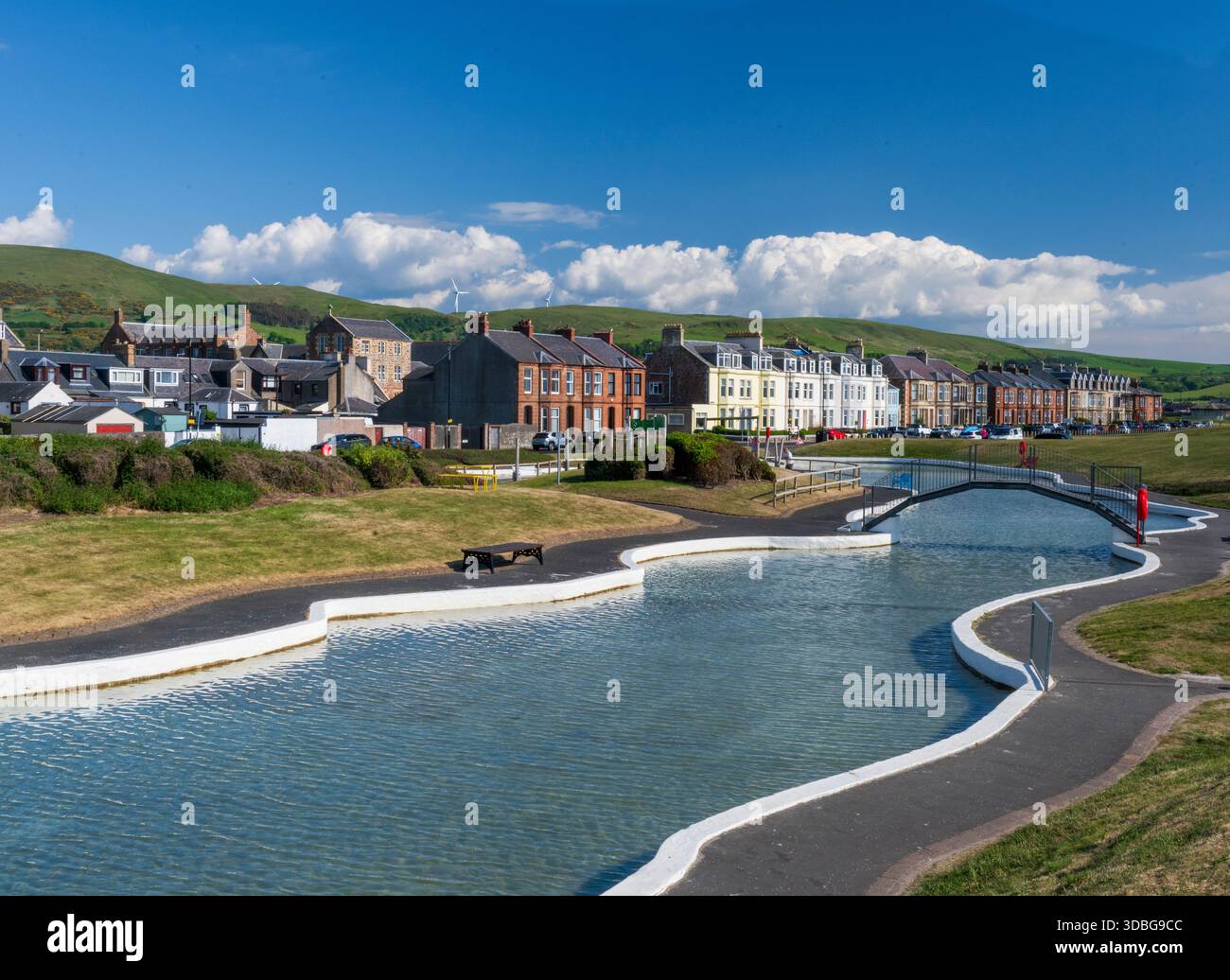 Scenario soleggiato del parco acquatico Girvan Esplanade con un canale, una fila di case colorate e colline lontane. Foto Stock