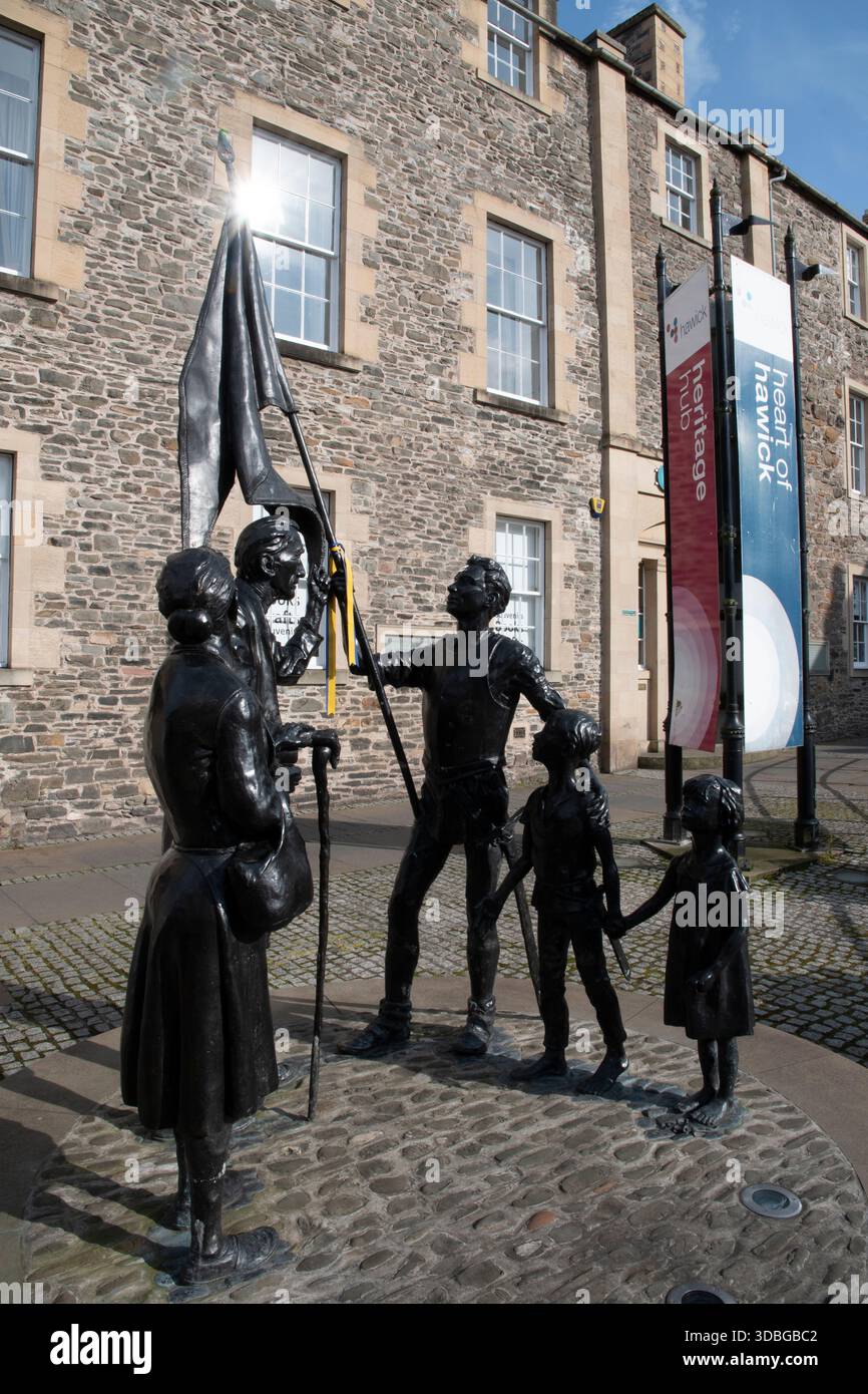 Scultura in bronzo di una famiglia e di adulti che innalzano una bandiera, situata accanto allo storico edificio in pietra di Hawick e alle moderne bandiere. Foto Stock
