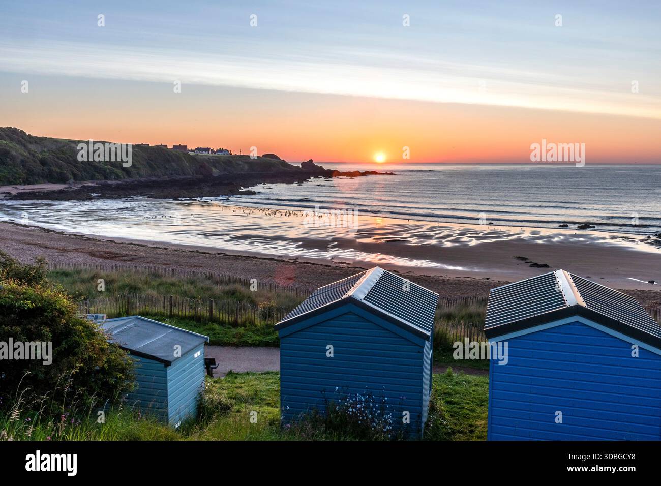 Tranquillo scenario costiero a Coldingham Bay con capanne blu sulla spiaggia, onde dolci e un vivace tramonto. Foto Stock
