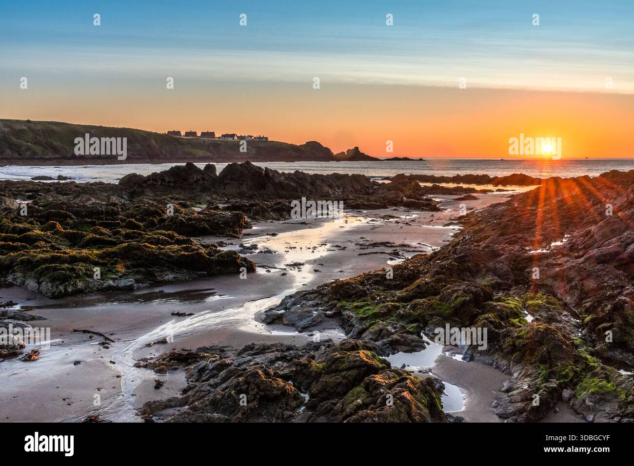 Tramonto costiero sulla costa rocciosa con muschio, acqua calma e cottage lontani lungo la baia. Foto Stock