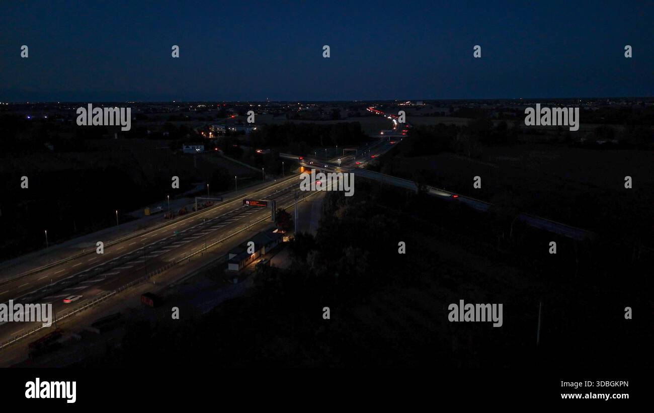 Vista aerea notturna dell'autostrada A21 con corsie illuminate, auto e camion in movimento attraverso un complesso interscambio e una piazza a pedaggio, mostrando un traffico intenso Foto Stock