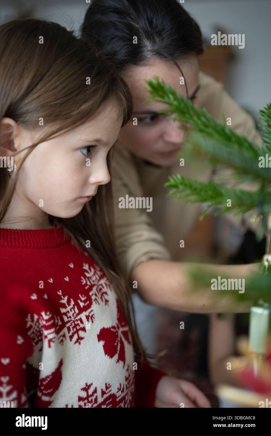 Ragazza in maglione rosso natalizio accanto alla madre che decora l'albero, accogliente vacanza in famiglia a casa Foto Stock