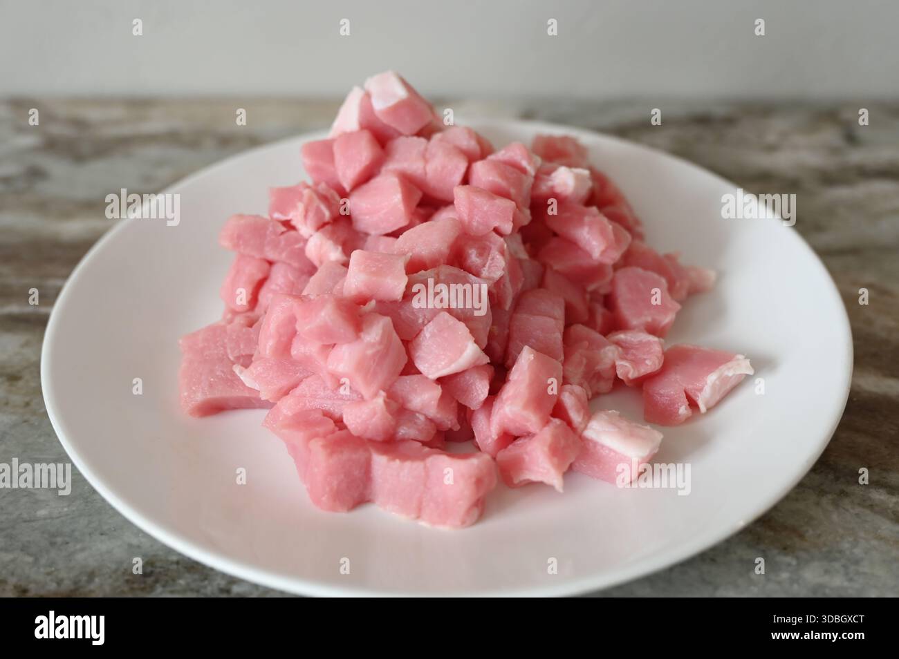 Pezzi di carne rosa a dadini impilati su un piatto bianco su un piano di lavoro della cucina, semplice concetto di preparazione degli alimenti, ingredienti crudi, stile rustico minimale sempre li Foto Stock