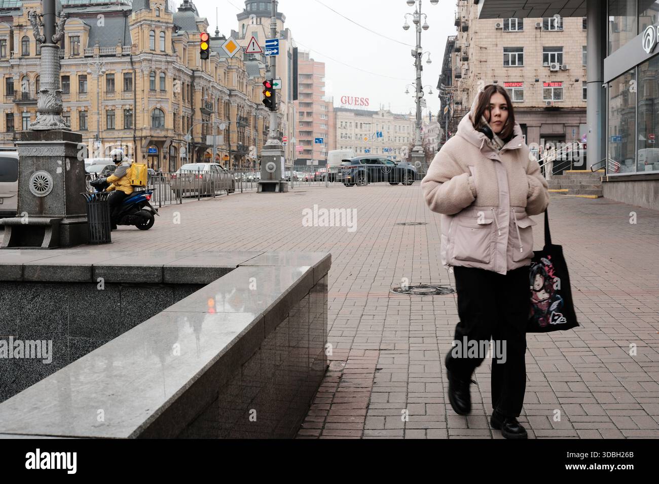 Una giovane donna cammina con sicurezza in una giacca accogliente attraverso una vivace piazza urbana. Kiev, Ucraina. 16 dicembre 2025. Foto Stock