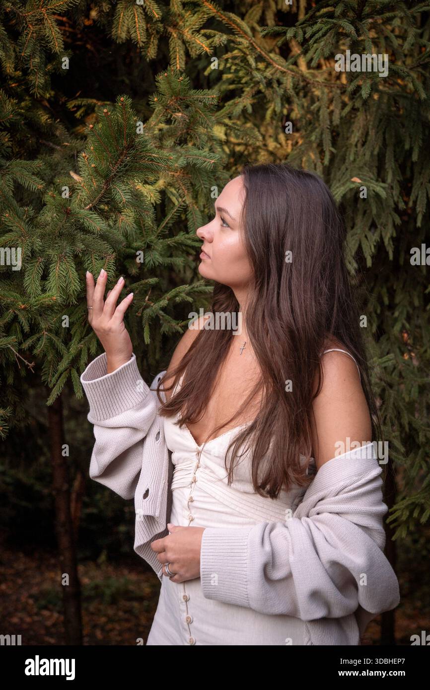 La giovane donna si trova vicino ai rami sempreverdi, toccando delicatamente gli aghi di pino, luce del giorno soffusa, toni verdi profondi, umore contemplativo Foto Stock