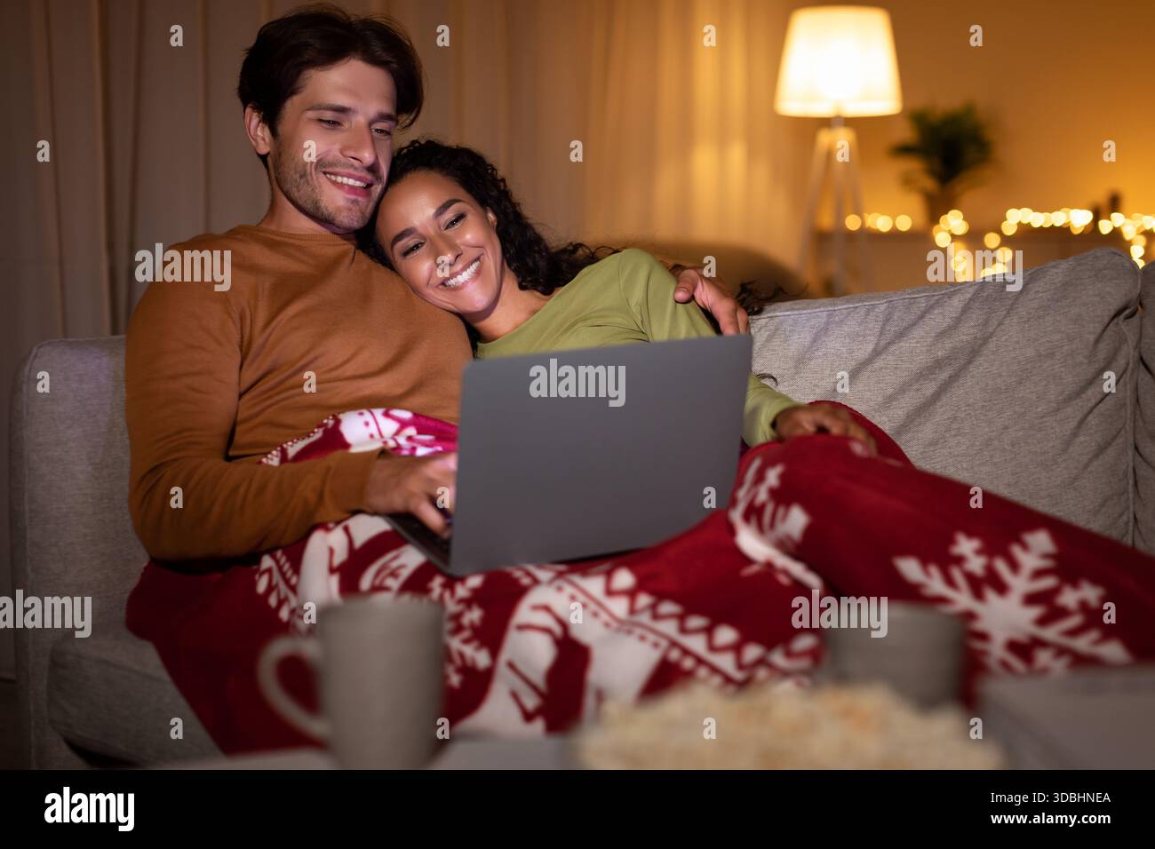 Le coppie si godono un'accogliente serata di film natalizi a casa mentre sono sdraiate sotto una coperta calda Foto Stock