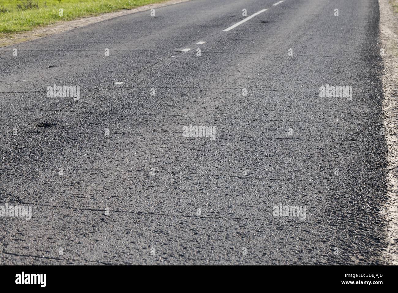 Una tranquilla strada rurale si estende in lontananza, mostrando asfalto fresco, sottili crepe e un luminoso vergine erboso sul lato della strada. La scena evoca trav Foto Stock