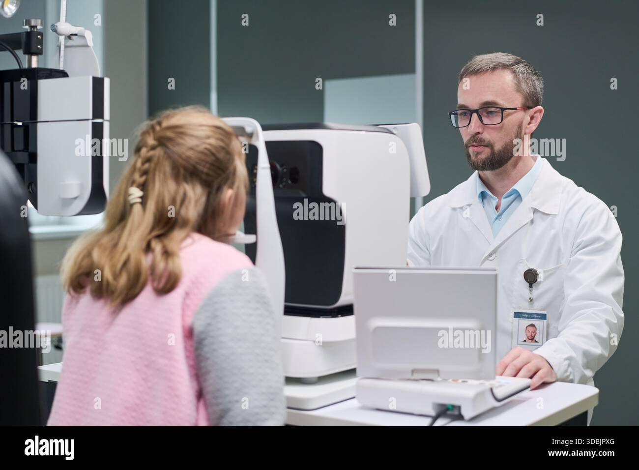 Uomo di mezza età caucasico optometrista che conduce l'esame oculare per la bambina caucasica utilizzando moderne attrezzature oftalmiche in clinica medica, entrambi concentrati sul processo diagnostico Foto Stock