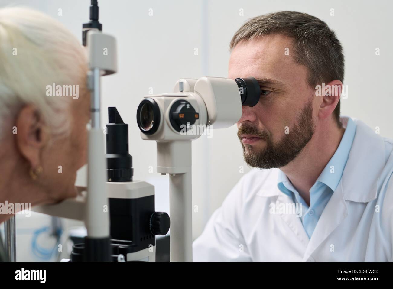 Uomo di mezza età caucasico che esegue l'esame oculistico su donna caucasica anziana utilizzando attrezzature oftalmologia in clinica medica, concentrandosi su pazienti valutazione della salute degli occhi Foto Stock