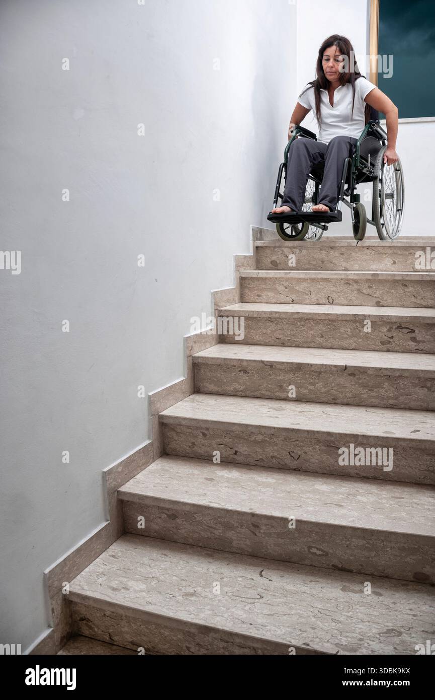 Una donna che utilizza una sedia a rotelle è posizionata in cima a una scala all'interno di un edificio pubblico o istituzionale. L'immagine mette in risalto l'architettura di barri Foto Stock