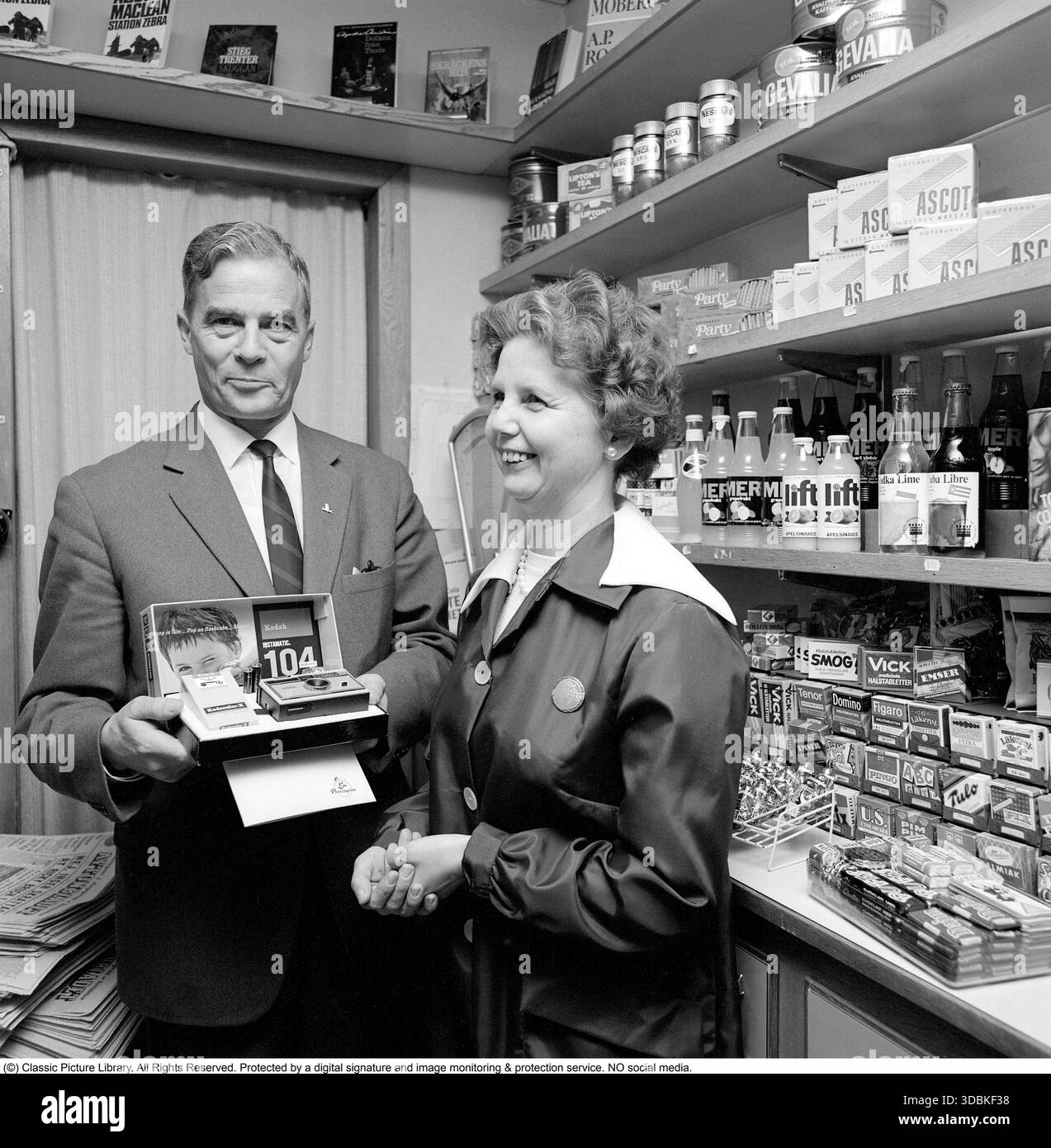 La Kodak Instamatic 104, introdotta nel 1965 e venduta fino alla fine degli anni '1960, era una fotocamera point-and-shoot accessibile e facile da usare che ha contribuito a rendere popolare la fotografia istantanea. Ha utilizzato l'innovativa cartuccia di pellicola 126 di Kodak, che ha consentito un semplice caricamento drop-in, senza la necessità di fastidiose infilature, rendendola ideale per utenti occasionali e famiglie. Con un obiettivo a messa a fuoco fissa, una presa per cubo flash integrato e simboli meteo per le impostazioni di esposizione, la serie Instamatic ha venduto oltre 50 milioni di unità in tutto il mondo, democratizzando la fotografia negli anni sessanta. Nella foto una dipendente donna in un piccolo negozio che riceve un Foto Stock